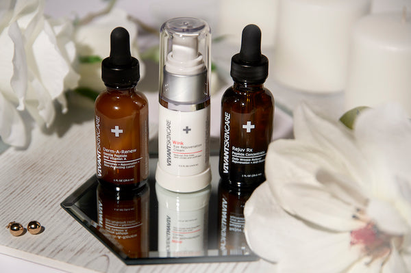 Two skin care serums and one eye cream over mirror and surrounded with flowers and candles. 