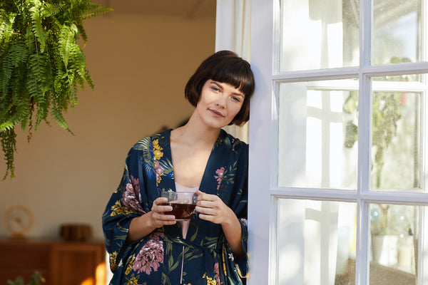 Portrait of a young woman in a robe leaning against her patio door in the morning and drinking a cup of tea