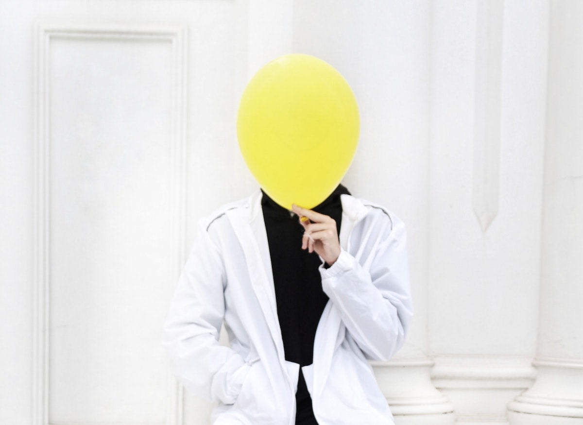 Man with a white jacket sitting with one hand inside a pocket while the other one holds a yellow ballon that covers his face.