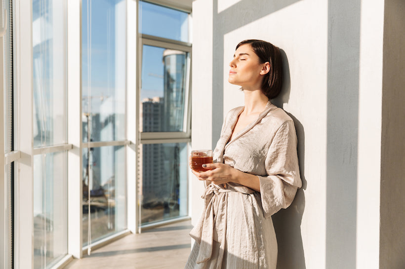 Elegant woman with short dark hair in housecoat holding glass of tea and enjoying sunny weather, while standing near window in posh apartment