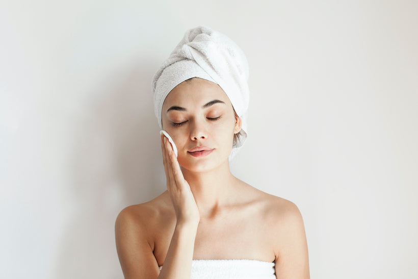 woman touching face with cotton pad, towel wrapping her hair