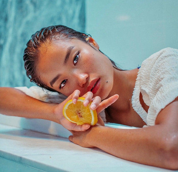 Asian looking woman leaning on a marble surface, with wet hair, and holding a orange slice while looking directly to the camera.  