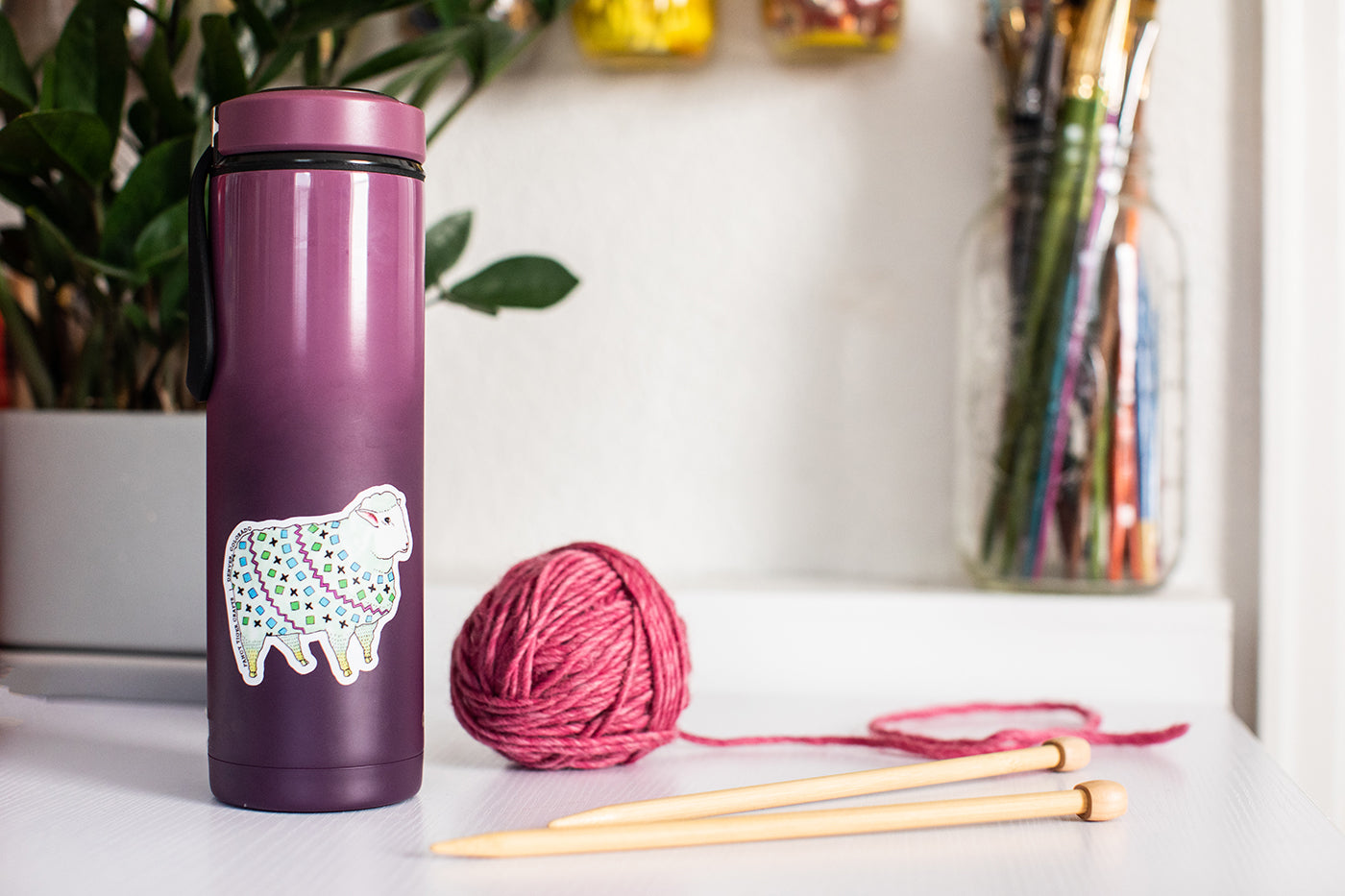 This is an image of a purple water bottle with a sheep sticker on the side of it and a ball of yarn and knitting needles sitting beside it