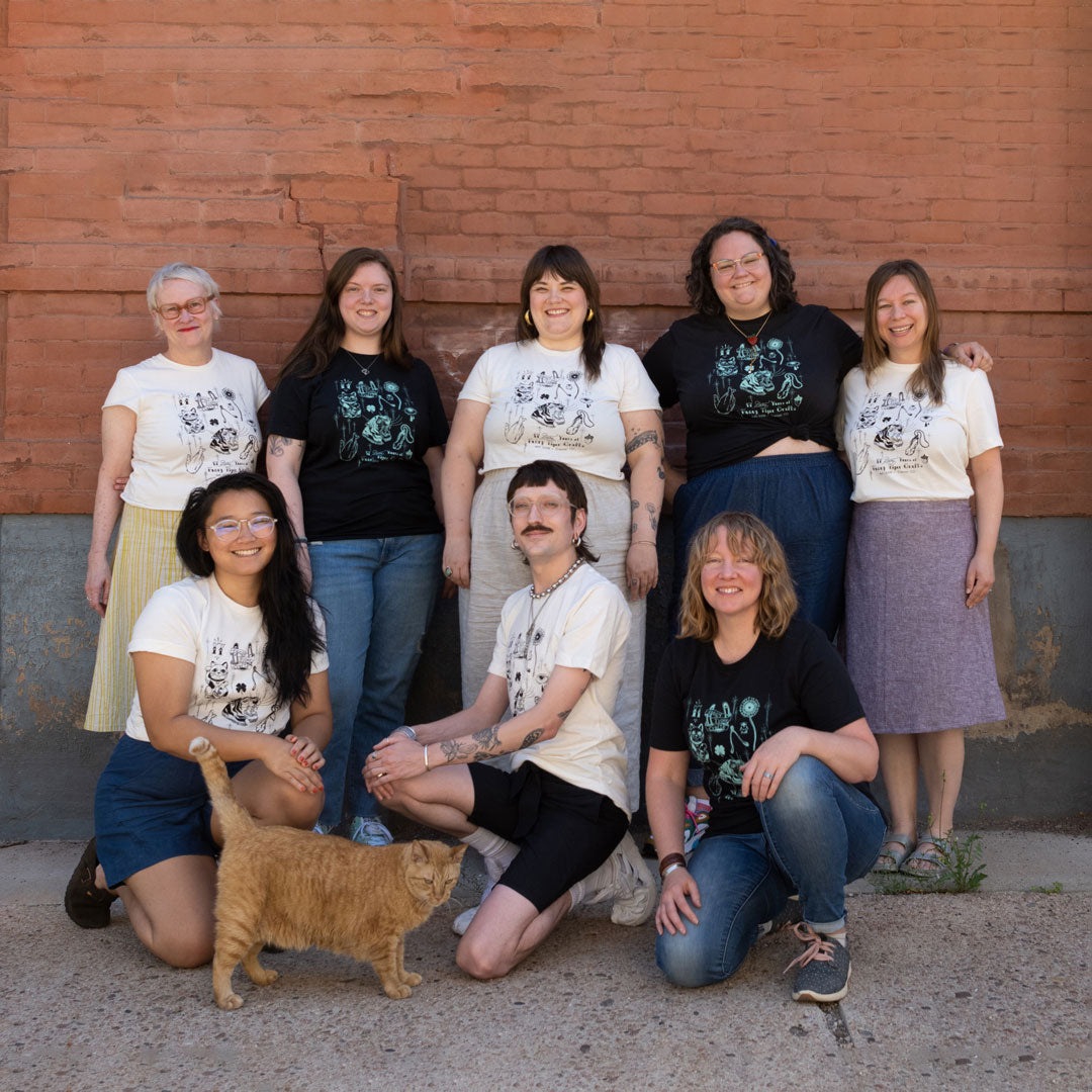 The Fancy Tiger Crafts staff stands in front of a brick way wearing their 17th Anniversary t-shirts either in black or white. An orange cat stands in front of them. 