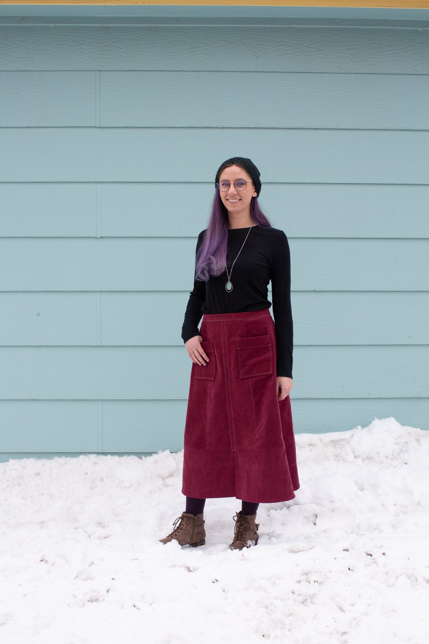 Aly standing in snow against a sea blue wall wearing a Reed Skirt in merlot corduroy and black long sleeve top.