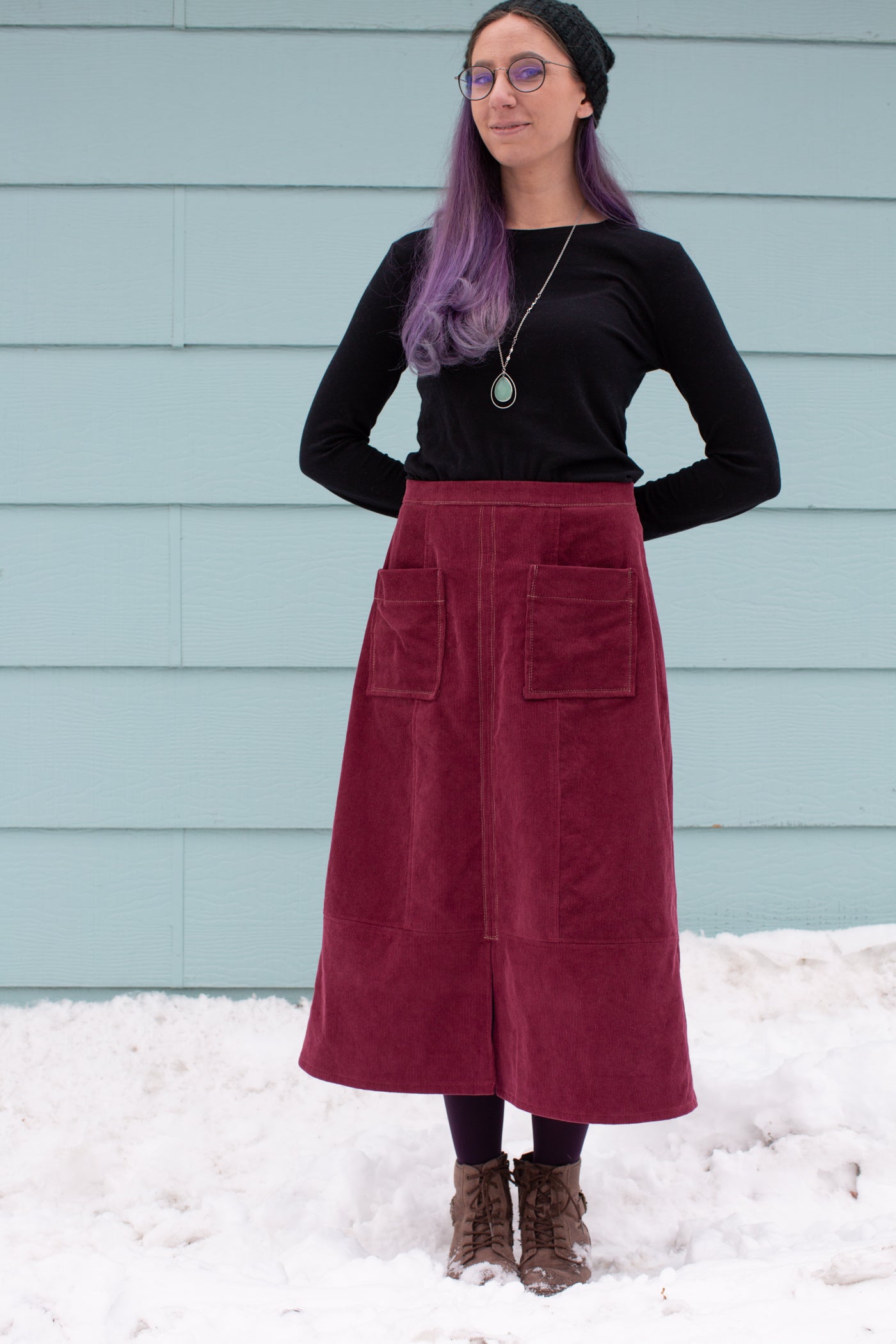 Aly standing in snow against a sea blue wall with her arms behind her back.  Aly is wearing a Robert Kaufman Corduroy skirt in merlot with a black long sleeve top.   