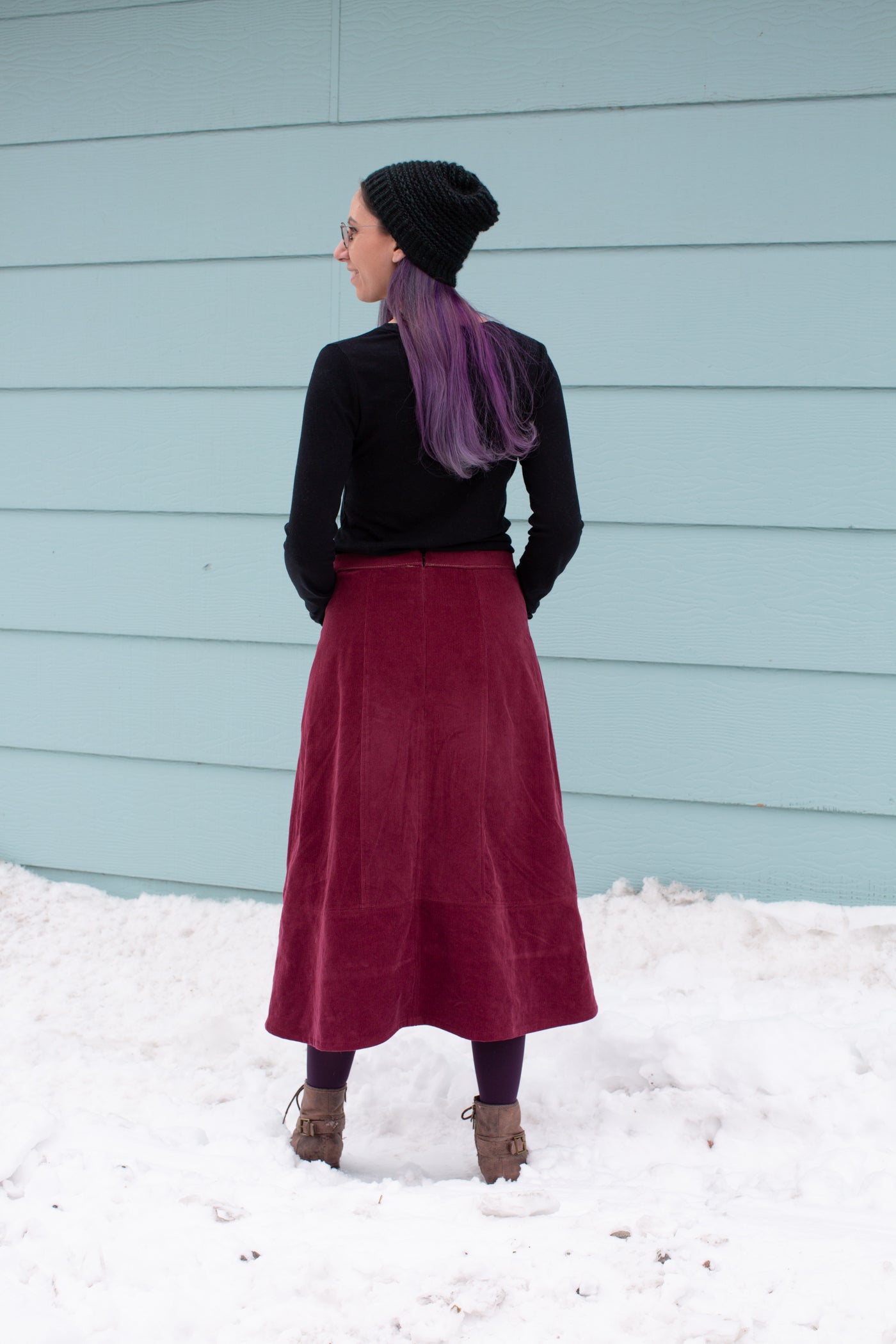 Aly is wearing a Robert Kaufman Corduroy skirt in merlot with a black long sleeve top.  Aly is standing in snow against a sea blue wall facing away from the camera, showing the back of the pattern.  