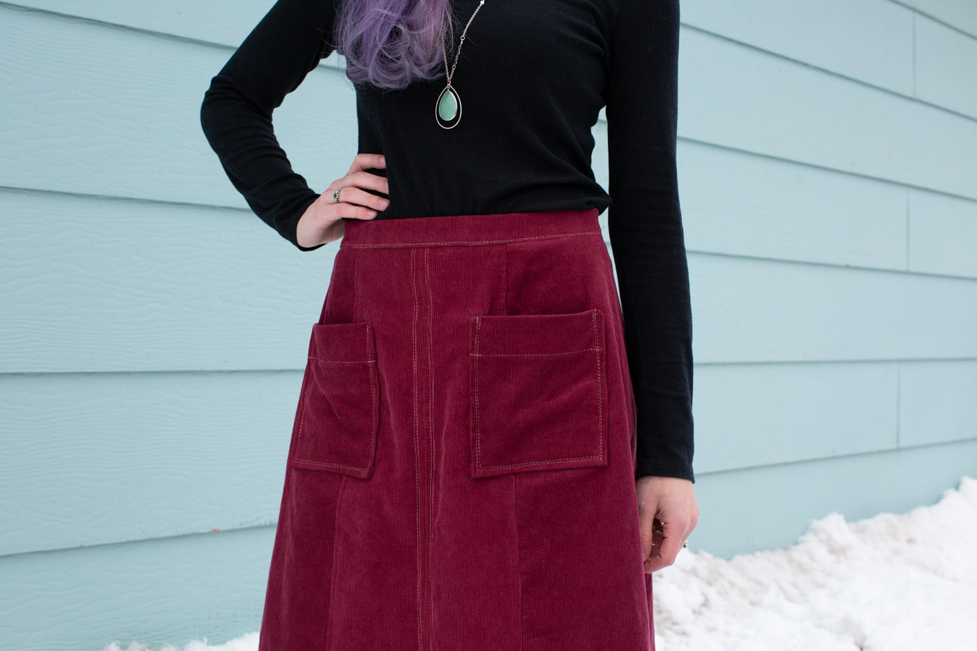 Aly standing in snow against a sea blue wall wearing a Reed Skirt in merlot corduroy and black long sleeve top. Close-up photograph of the the pockets with white stitching on the skirt..