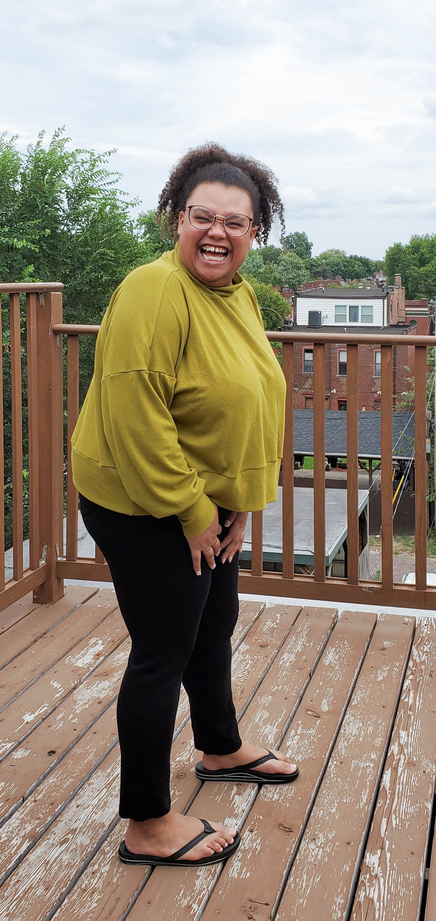 Women wearing a Hosta Tee long sleeve in Bamboo Cotton Stretch Rib in Chartreuse and black jeans.  Women is standing on an outdoor patio in the city with cloudy blue skies, sideways with right shoulder and face turned towards the camera.  Women is smiling and laughing..