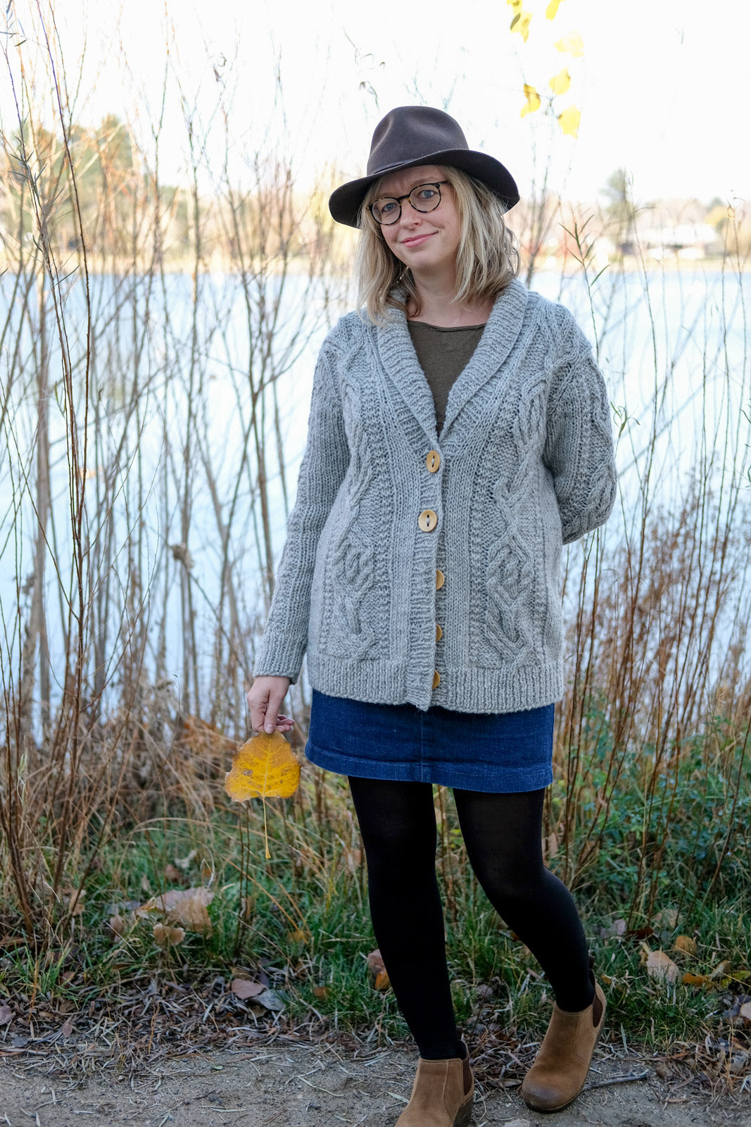 Amber by the lake holding a leaf in her Cabled Junegrass Sweater