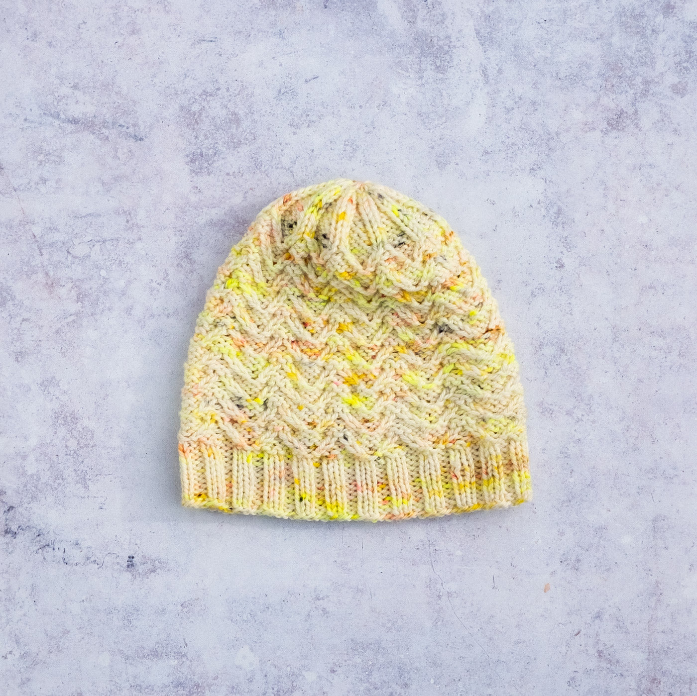 Handknit hat laying flat, it has textured stitches in white yarn speckled with neon yellow, gold and flecks of coral here and there. The crown of the hat has in interesting decreasing pattern, the texture of the stitches come together a little like a flower motif.