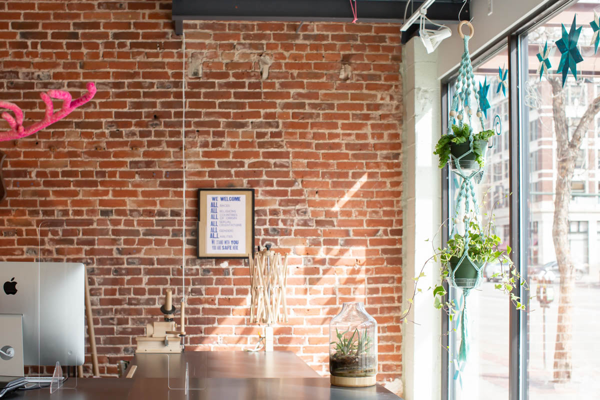 The two-tiered macramé hanger in mint green hangs in the bright front window of Fancy Tiger Crafts. to the left is the tidy cutting table with yarn swift where staff cut fabrics and prepare purchases. On the red brick wall behind the counter hangs a sign: "We welcome all races, all religions, all countries of origin, all sexual orientation, all genders, all abilities. We stand with you. You are safe here."