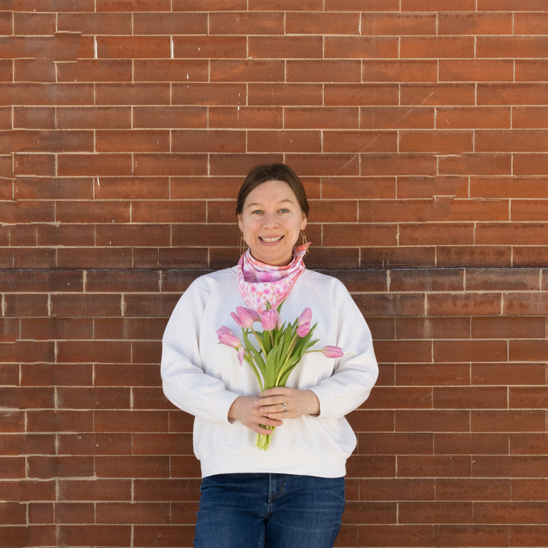 This is an image of Anna standing in a front of a brick wall. She has her Kaleidoscope scraf wrapped around her neck and she is holding a bouquet of pink tulip. Anna is also wearing a white sweater and blue jeans. s 