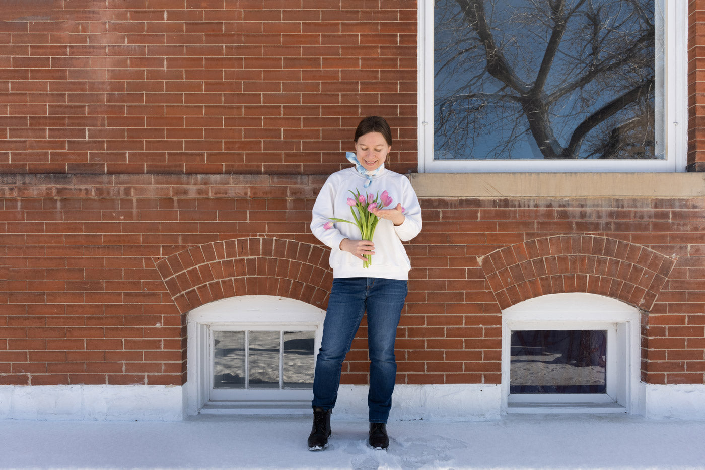 This an image of Anna wearing her dyed blue scarf around her neck. She is looking down at the bouquet of pink tulips she is holding while standing in front of a brick building. 