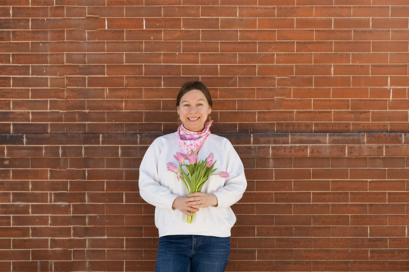 Anna stands in front of a brick wall. She is wearing a white sweater with a dyed pink bandana around her neck and holding a bouquet of tulips. 