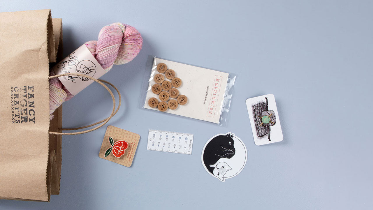 The top of a brown paper bag with the text "Fancy Tiger Crafts" stamped on it layed horizontal across a blue background. A pink skein of yarn, a flower pin, wooden buttons, cat sticker, and a pin of a ball of yarn are positioned to appear like they're overflowing out of the bag. 