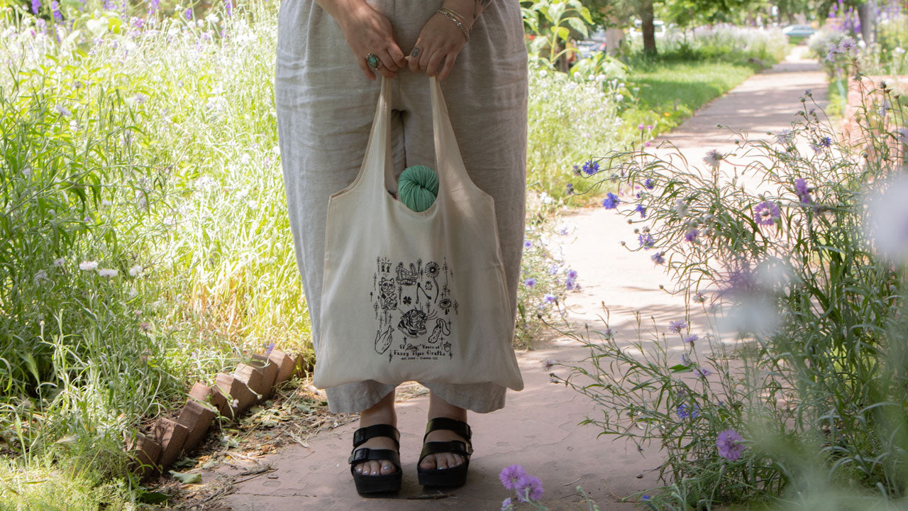 A limited edition 17 Anniversary tote bag is being held by a person wearing white pants and black platform sandals. A mint green skein of yarn peaks out of the bag. The person is standing on a path surrounded by flowers