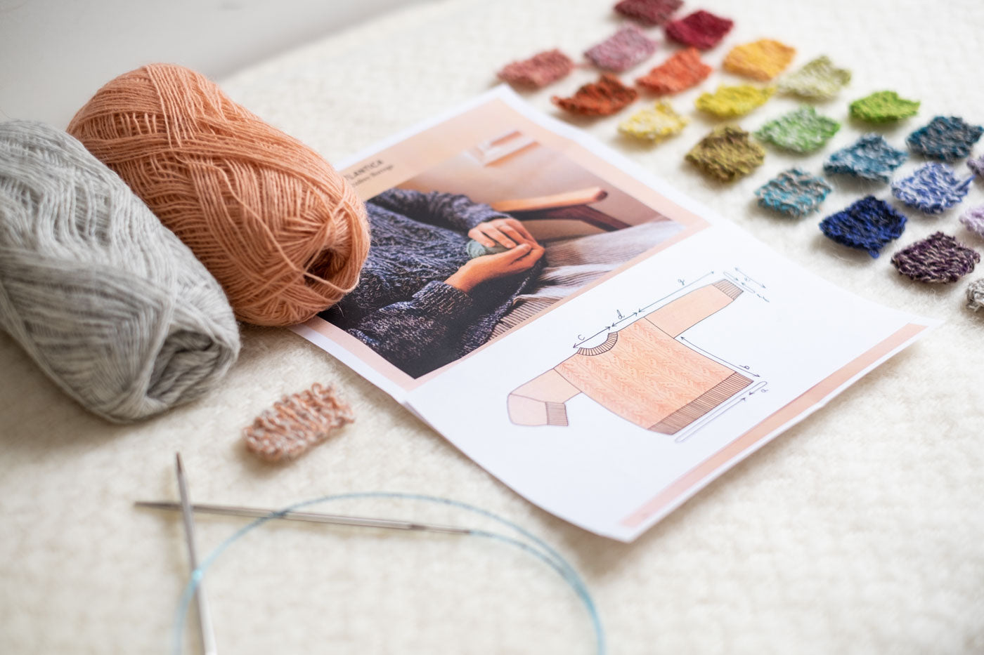 Two balls of yarn in gray and pale peach are displayed next to a pattern featuring a cable-knit sweater along with dozens of small knit swatches and knitting needles.