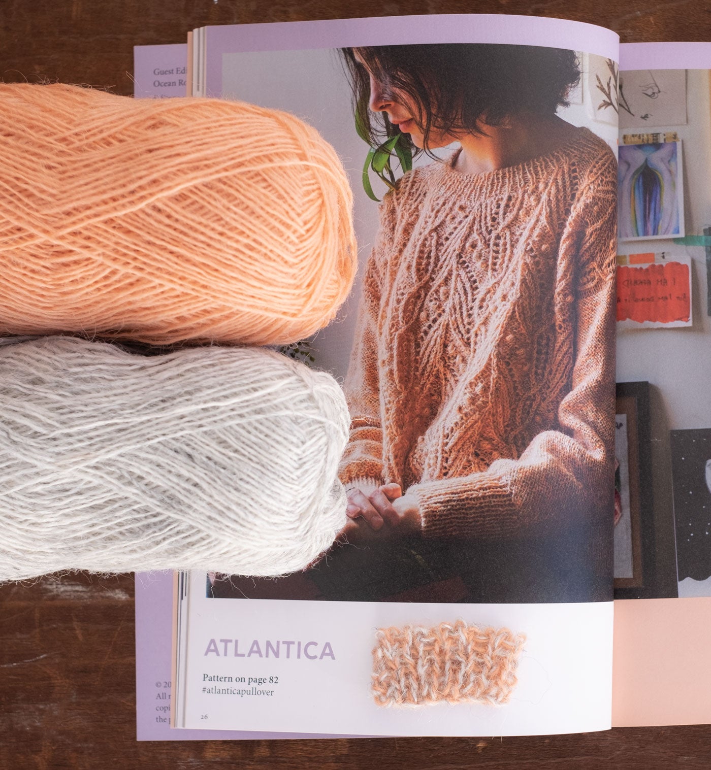 A magazine lays open to a page showing a person with chin-length curly hair wearing the Atlantica Pullover—a delicate lace and cable sweater with a slightly oversized fit. It is a pale, muted peachy color. Two balls of laceweight yarn are on top of the magazine, peeking into the frame of the picture. One is pale heathered grey and one is a warm pale peach. They are held together to created the muted marl fabric. 