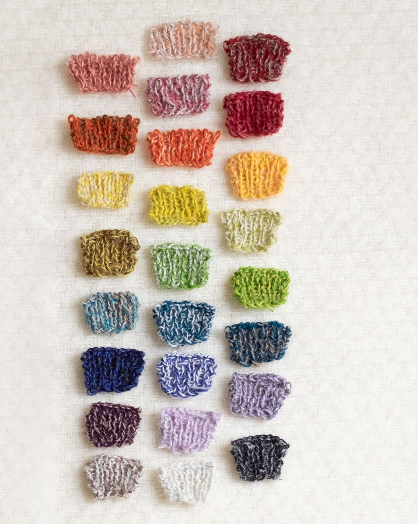 25 tiny knitting swatches in a rainbow of various combinations of two-color marls are laid out neatly on a soft, fuzzy, white woven wool background.