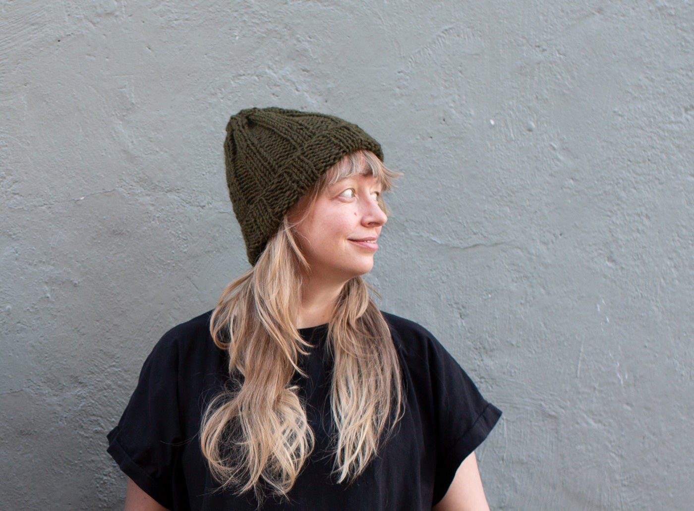 Amber stands in front of a grey wall, looking to the side, and wearing a cozy olive-hued, chunky knit beanie..