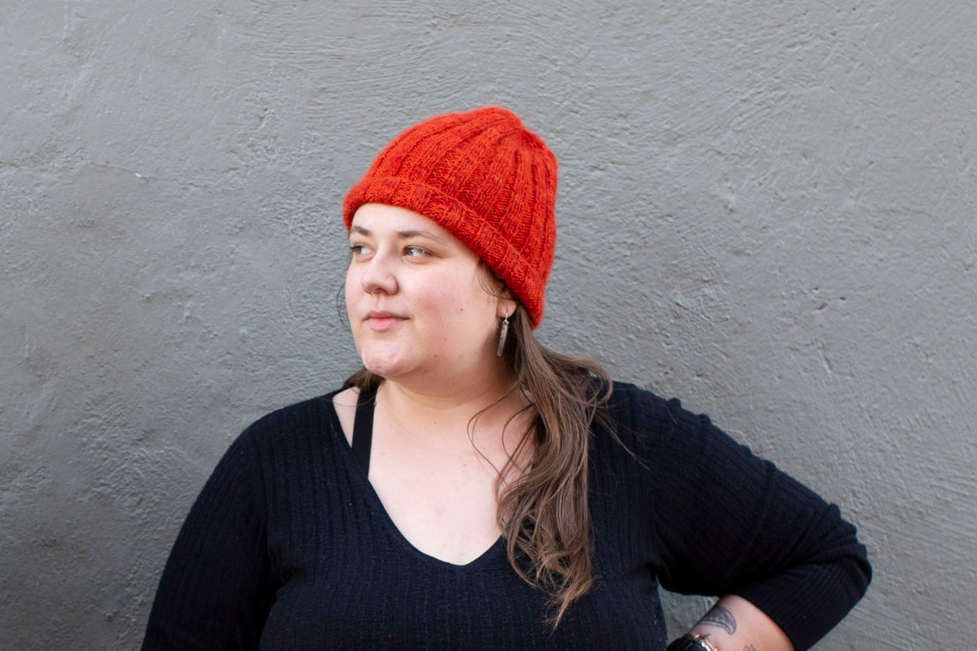 Marta stands in front of a grey wall, looking to the side, and wearing a vibrant, flame red-orange, fuzzy ribbed beanie.