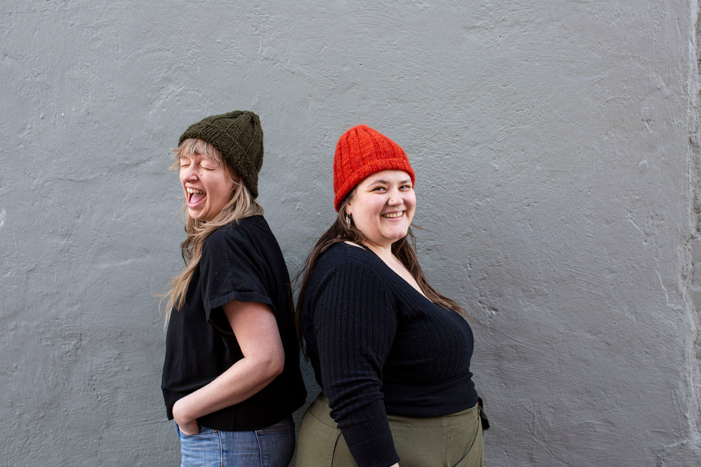 Amber and Marta stand back to back against a grey brick wall. Amber makes a silly face and wears a bulky, olive-hued ribbed hat. Marta has a laughing smile and wears a finer knit, flame red-orange ribbed hat, with a fuzzy texture. 