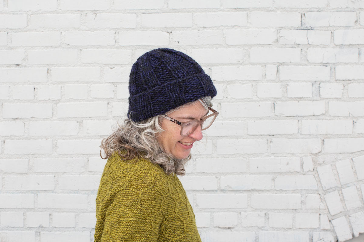 Jaime looks down, smiling. She wears a dark blue-purple and black marled ribbed beanie, and a sap-green handknit sweater.