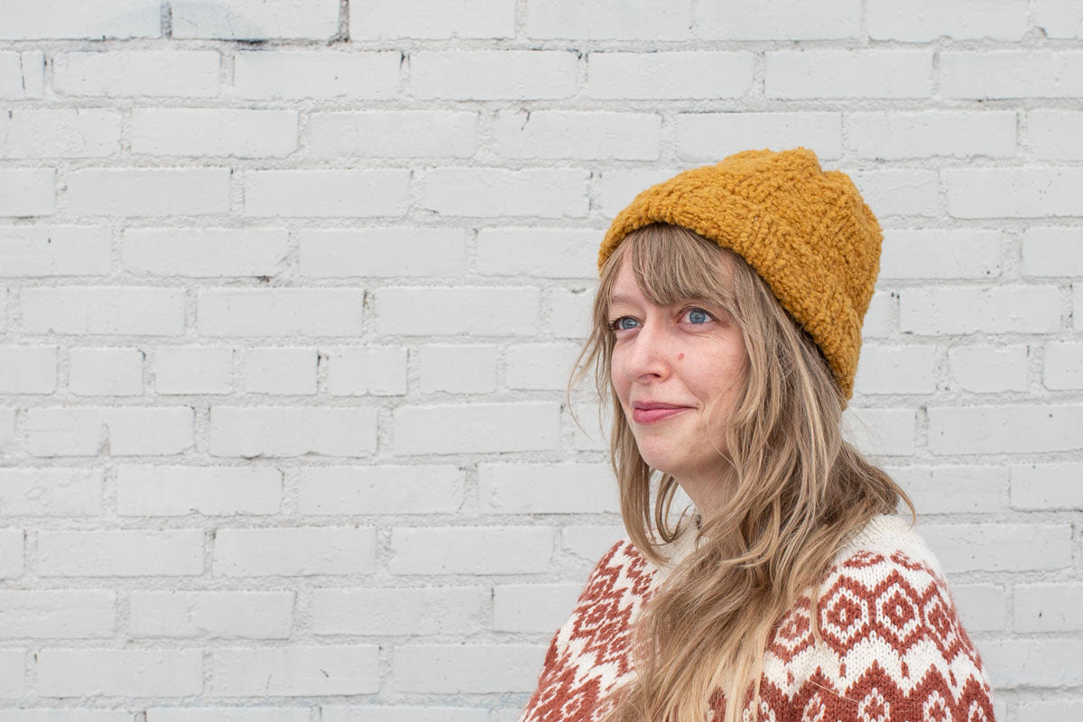 A close up of Amber wearing a handknit ribbed beanie—a bulky boucle yarn gives the hat a soft, sherpa-like texture. She wears a coppery brown and white colorwork yoked sweater. 