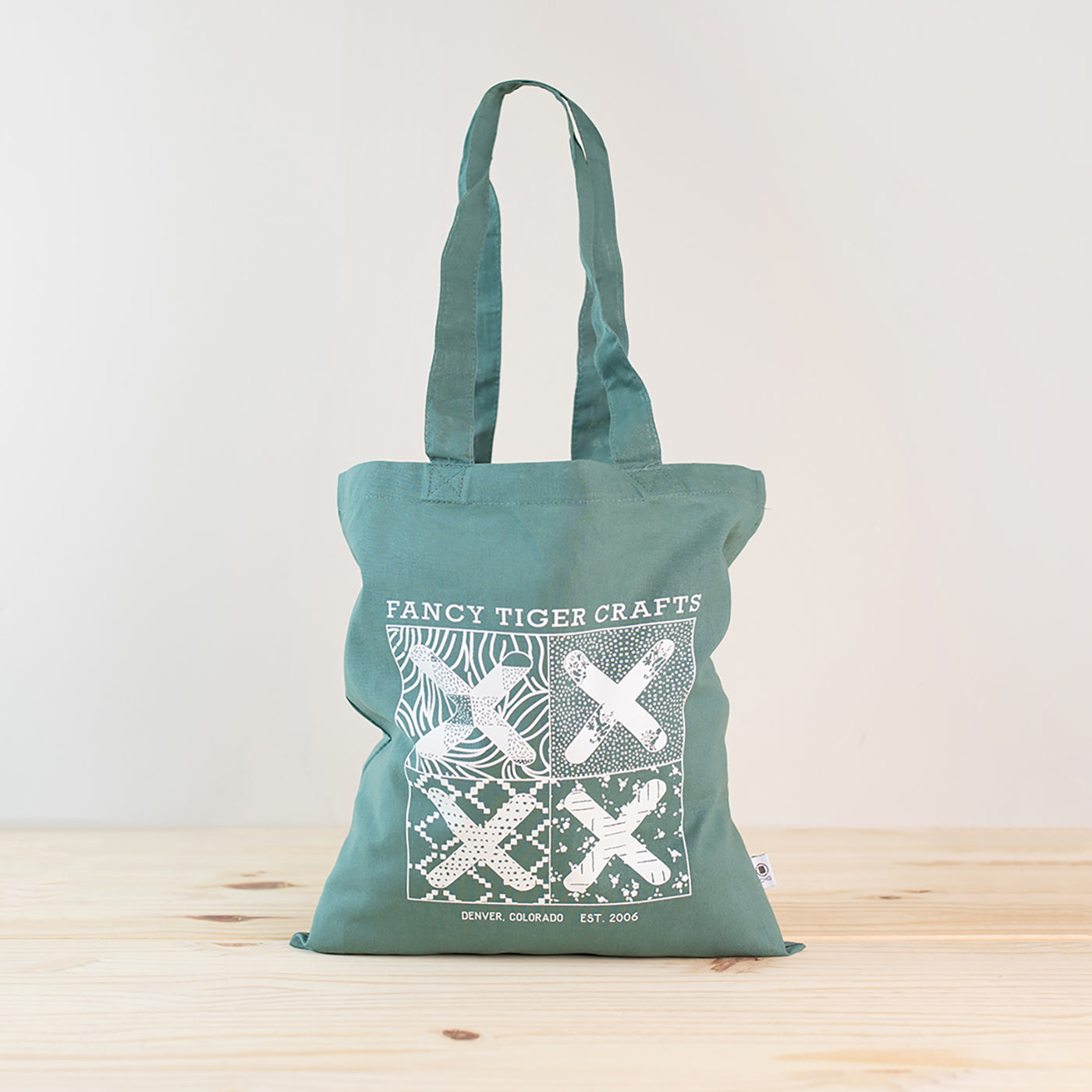 This is an image of a reusuable tote bag with the Fancy Tiger Crafts logo on the front of it sitting in front of a white wall and on a wood floor