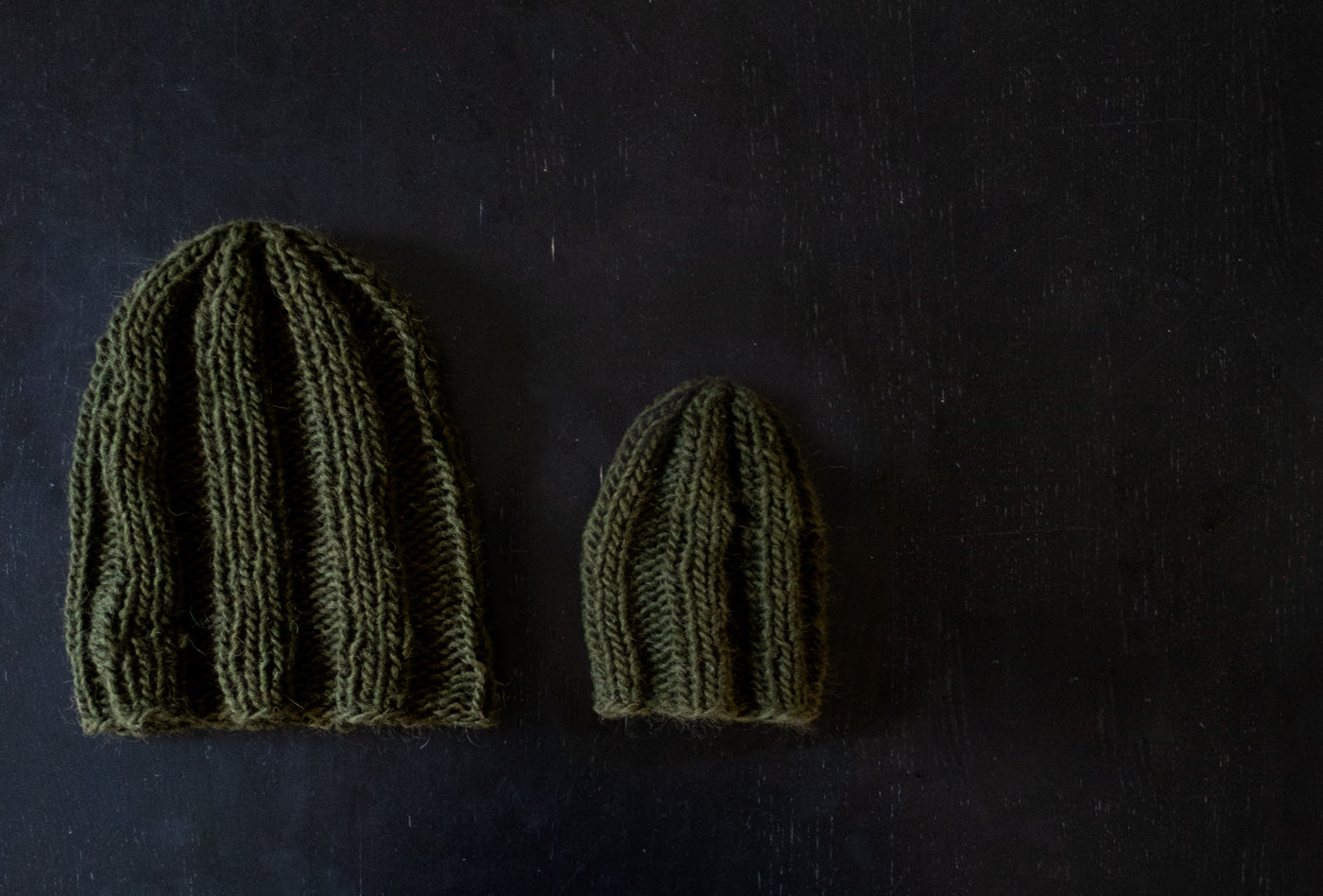 Two olive-hued, chunky knit hats lay on a dark wood background. The hat in the center is much smaller than the adult hat to the left of the photo. Adorably small, it is sized for a baby or toddler. 