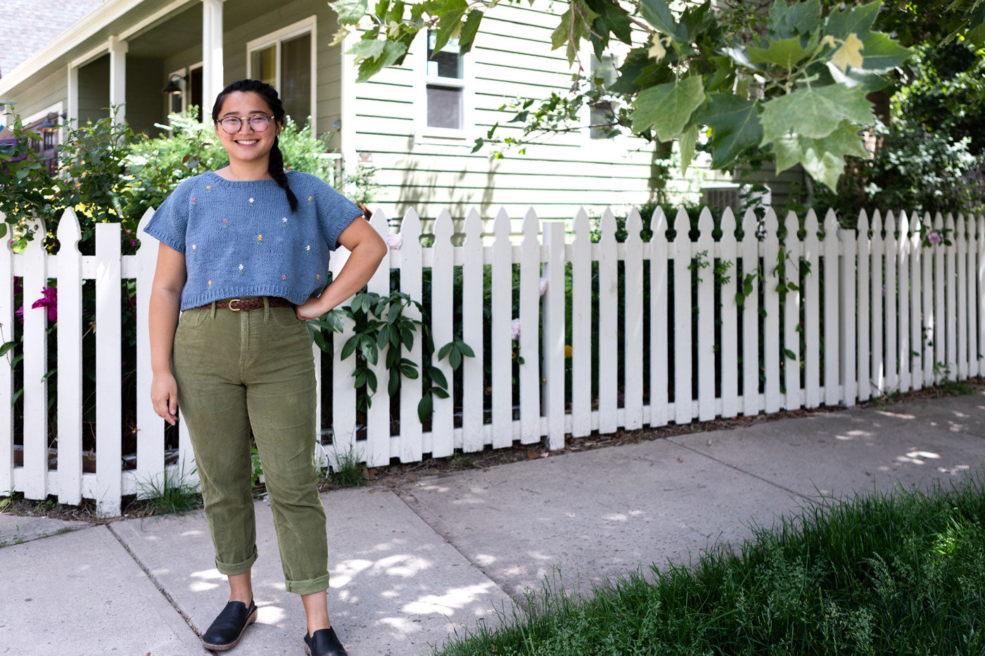 Bethany stands in front a white picket fence. She is wearing a blue knit top with embroidered flowers and wearing green pants.