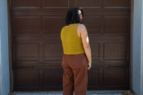 Bethany stands facing a brown garage door with a hand knit mustard yellow tank top