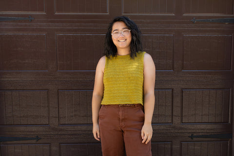 Bethany stands in front of a brown garage door with a hand knit mustard yellow tank top