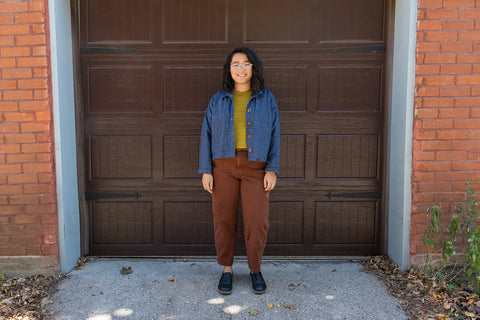 Bethany stands in front of a brown garage door with a hand knit mustard yellow tank top and a light weight denim jacket