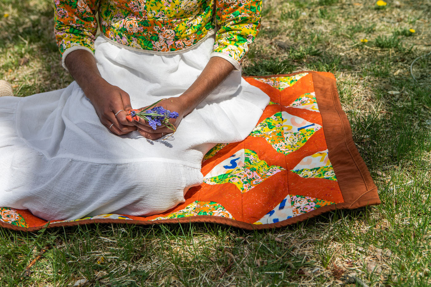 This is a close up image of Beza sitting on her Charming Xs quilt, which is laid out on a grassy lawn. She holds a small bouquet of purple flowers in her hands. 