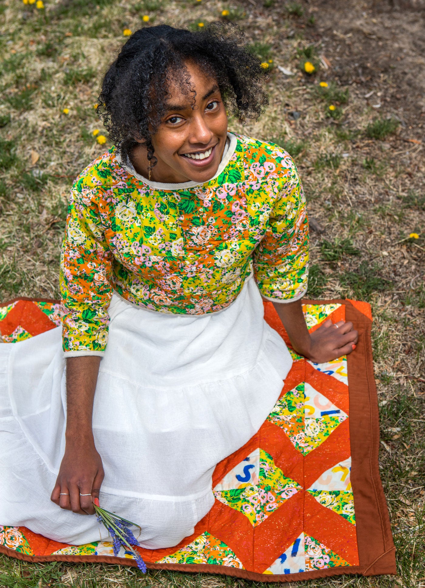 Beza sits on her Charming X's quilt that is laid out on a grassy lawn. She is looking up at the camera with a wide smile. 