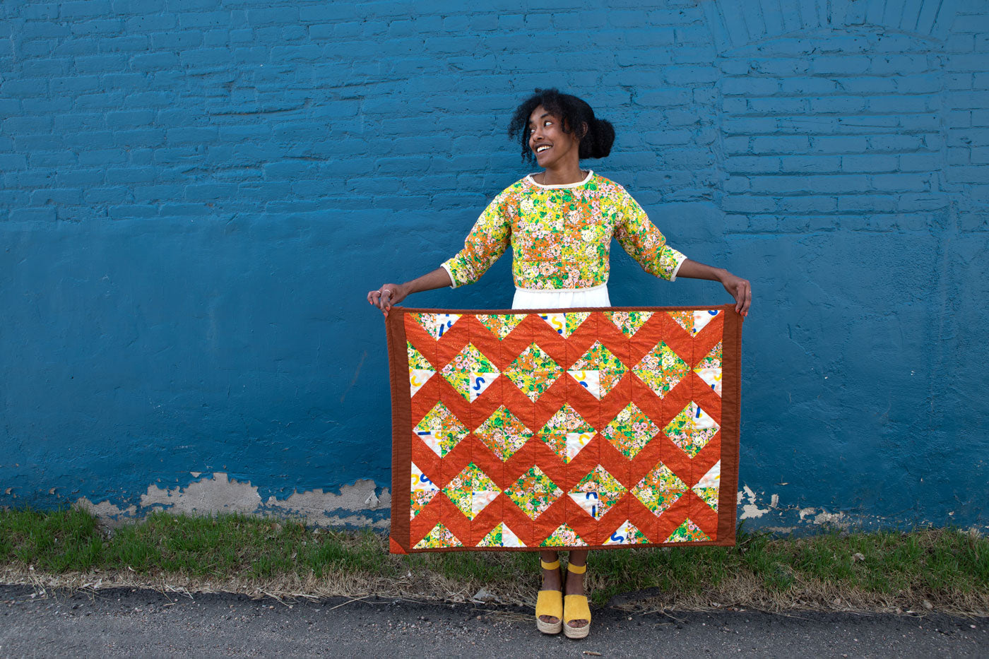 Beza stands in front of a blue wall holding her Charming Xs quilt. Her face is looking to the side with a wide smile, she is wearing a colorful quilted top and bright yellow shoes. 