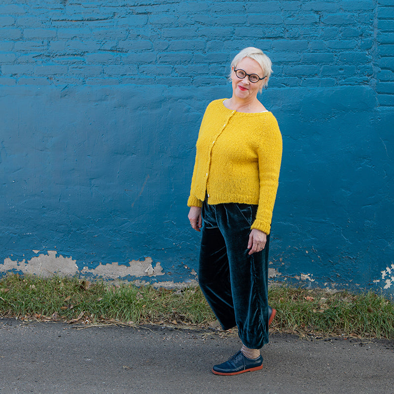 This is an image of Christina wearing her Emerson Pants by True Bias in Silk/Rayon Velvet in Peacock Blue. She is wearing a yellow knit cardigan and standing in front of a blue wall. 