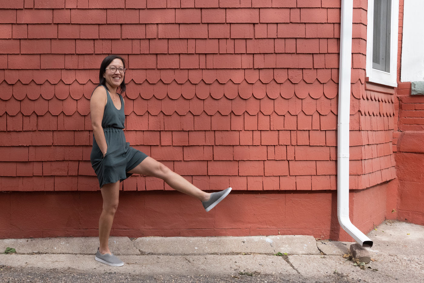 Jenn stands sideways next to a red wall, she has one leg kicked out in front of her with her hands in the front of her Nova Jumpsuit pockets