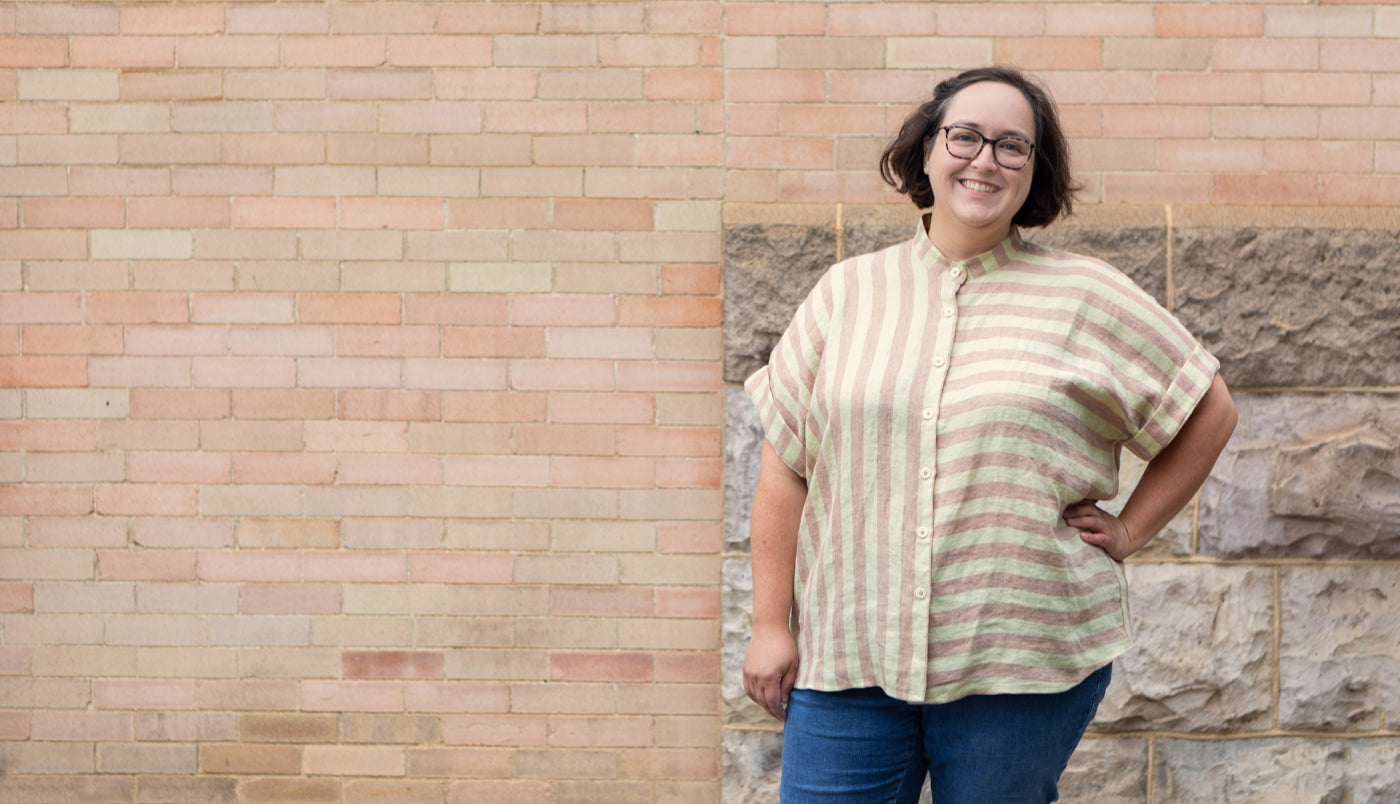 This is an image of Caitlin wearing a pink and white striped Merchant and Mills  linen button down shirt by The Assembly Line standing in front of a brick wall. 