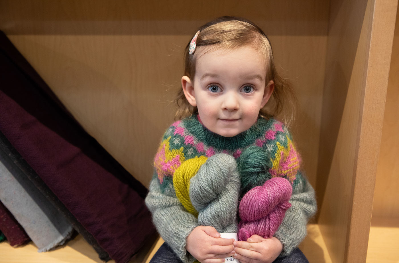 Caitlin's daughter sitting next to some fabric bolts on a shelf, she is holding a bundle of Heirloom Romney skeins while wearing her Mini Kordy sweater.