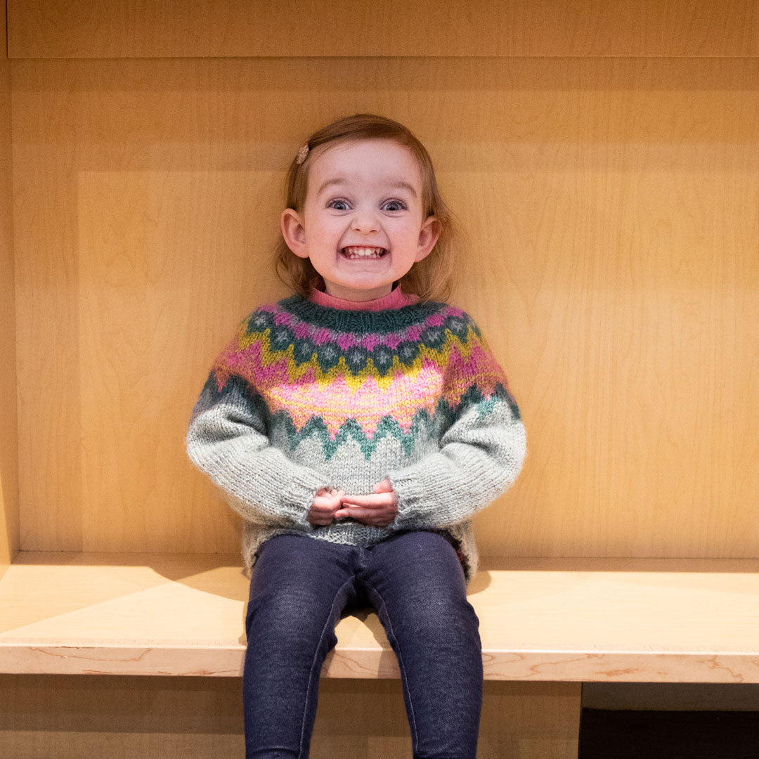 Caitlin's daughter sits on an empty bottom shelf wearing her Mini Kordy Heirloom Sweater sweater