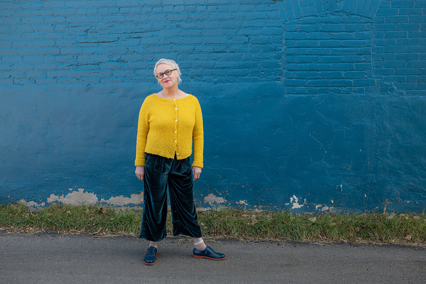 This is an image of Christina wearing her Emerson Pants by True Bias in Silk/Rayon Velvet in Peacock Blue. She is wearing a yellow knit cardigan and standing in front of a blue wall. d