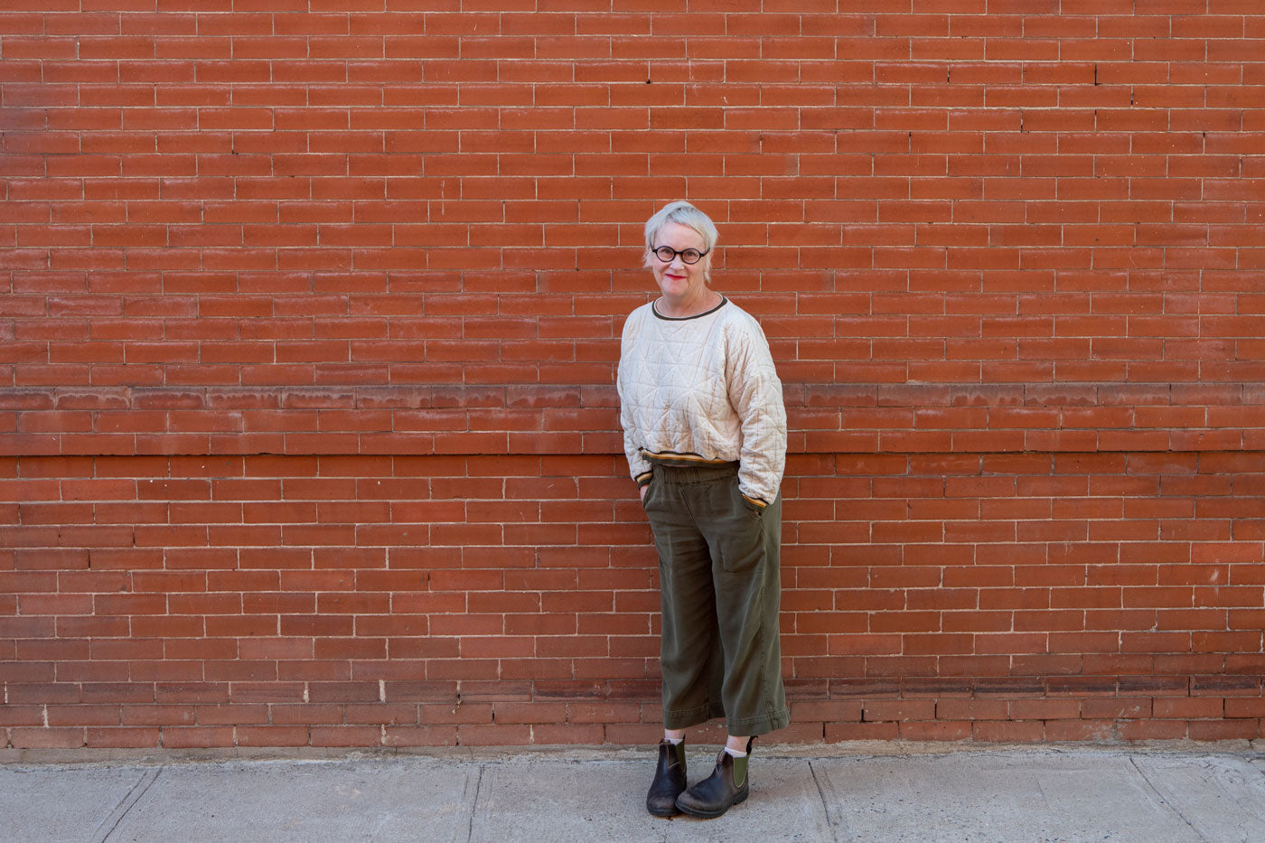 Christina stands in front of a brick wall. She is wearing a white sweater with green pants. 