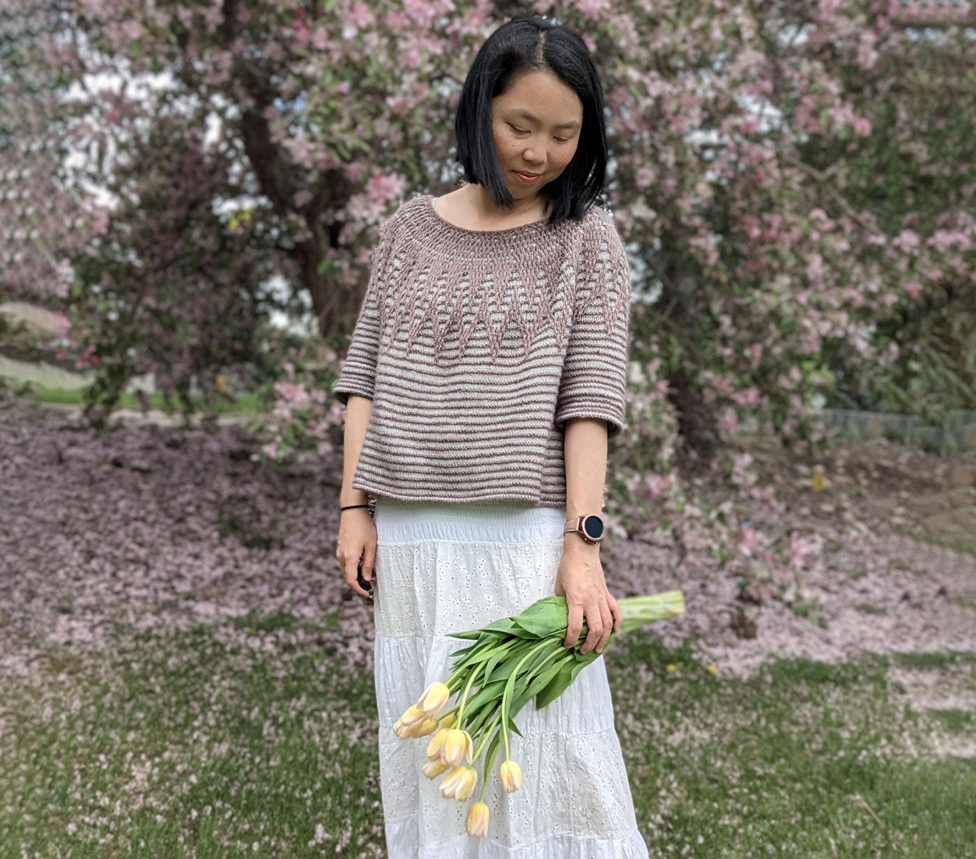 Connie looks down at the bouquet of tulips she's holding. She is wearing her Aubade pullover and long white skirt. She stands in front of a pink cherry tree.