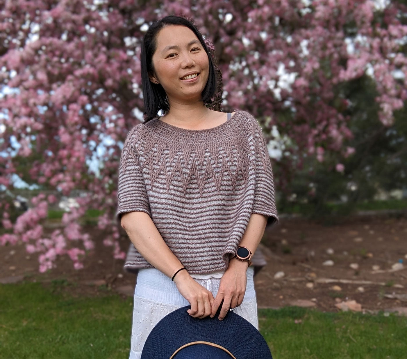 Connie smiles at the camera while wearing her Aubade sweater and wearing a long white skirt. She stands in front of a cherry tree and is holding a dark navy wide brim hat.