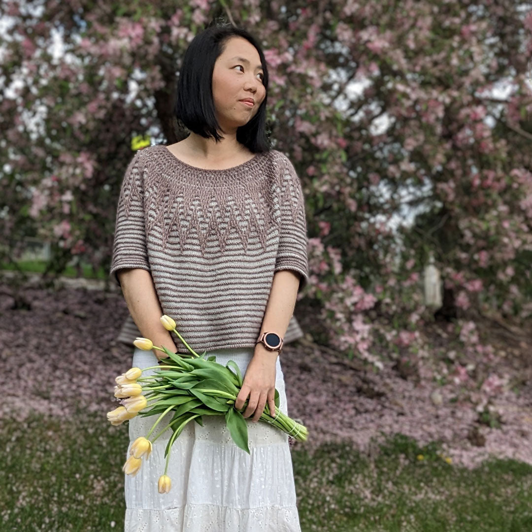 Connie looks off into the distance wearing her Aubade pullover and long white skirt. She stands in front of a pink cherry tree and is holding a bouquet of tulips. 