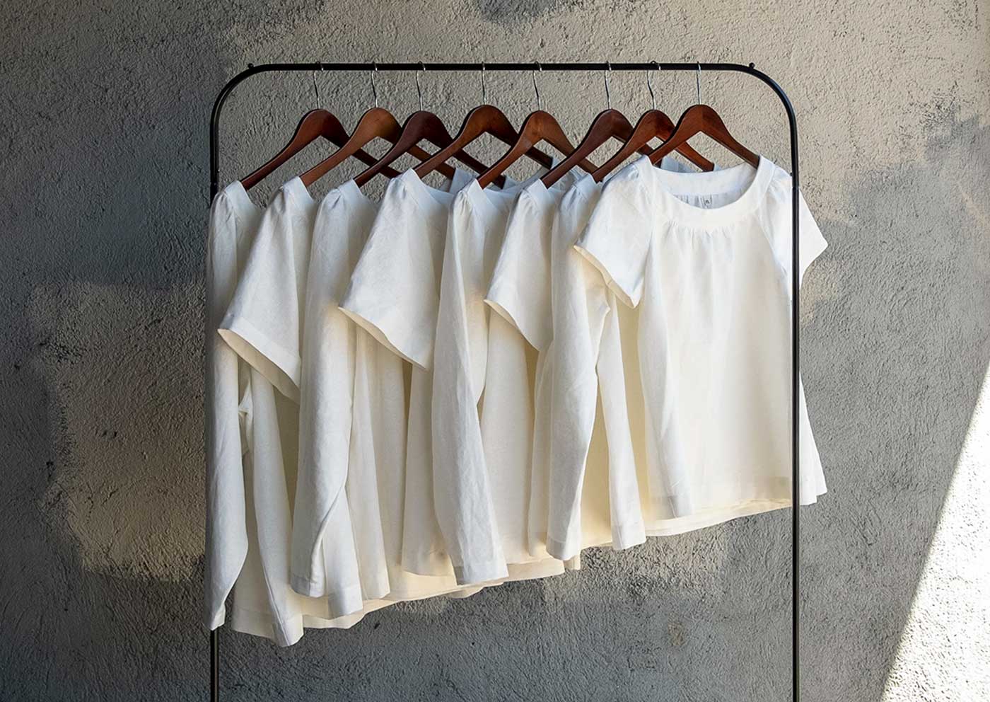 Eight solid white linen Cordyline tops in a range of sizes hanging on a rack. 