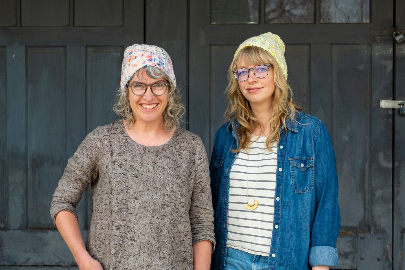 Jaime and Amber standing together, both wear handknits hats that are white with neon speckles. 